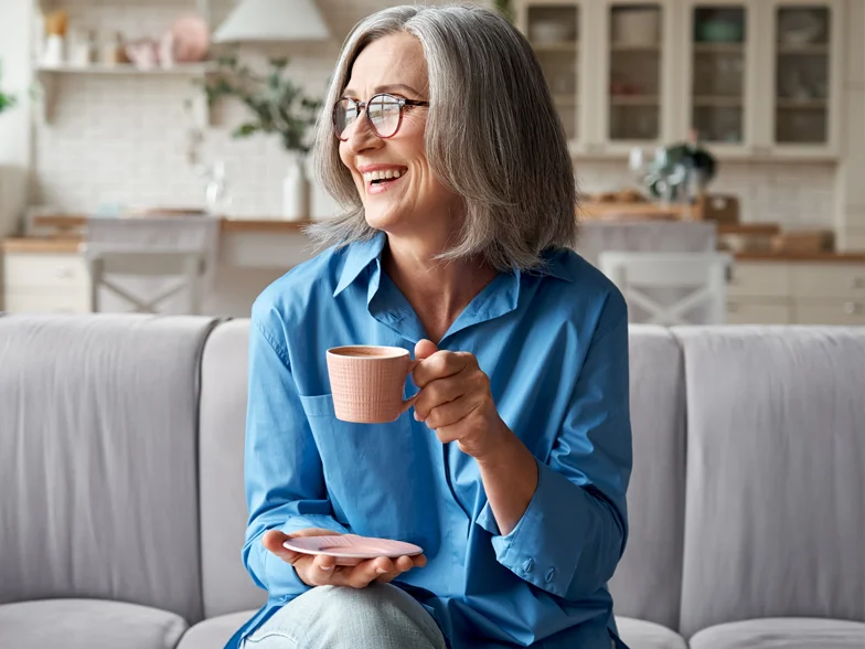 A happy and beautiful, relaxed, mature, grey-haired woman drinking coffee and relaxing on a sofa in a modern living room.